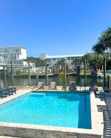 Waterfront swimming pool with blue Adirondack chairs, dock pilings, and palm trees overlooking canal